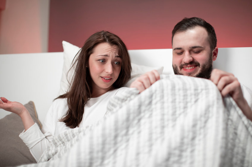 happy smiling man in bed looking under blanket at penis, wife looks surprised